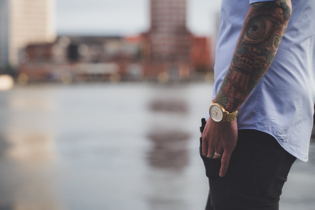 Close-up of a tattooed arm with watch against blurred city backdrop, capturing modern urban style.